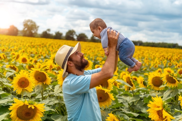 Portrait of a father in a blue shirt and a straw hat and his baby having fun in a field of sunflowers, summer lifestyle