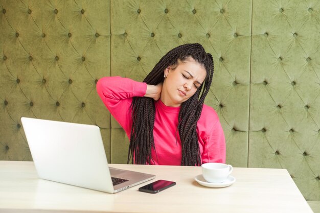 Portrait of feel bad tired young businesswoman with black dreadlocks in pink blouse is sitting in cafe and holding neck with hand, have neck stiff, making massage. Indoor, health care