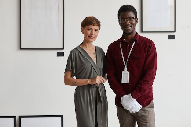 Portrait of female art gallery manager posing with African-American worker while standing against white wall and smiling at camera, 