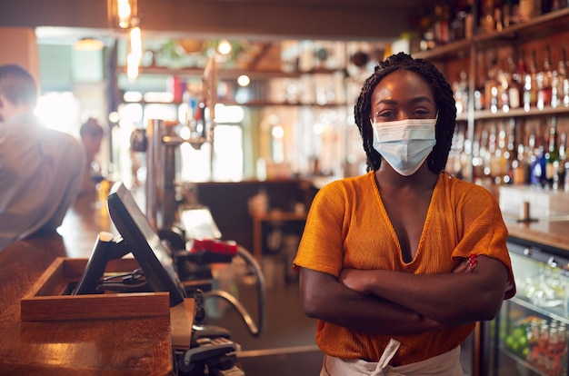 Portrait of female bar worker wearing face mask during health\
pandemic standing behind counter