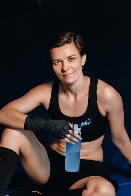 Портрет женщины-боксера в красных перчатках в тренажерном зале после тренировки с бутылкой воды.