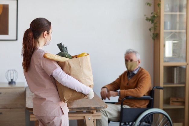 Ritratto di badante femminile che tiene in mano una borsa con generi alimentari mentre consegna cibo a un uomo anziano in sedia a rotelle, copia spazio