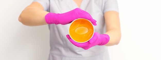 Portrait of a female caucasian beautician holding a jar of sugar paste for sugaring wearing pink gloves on white background