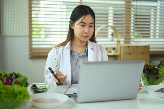 백색 코트를 입은 여성 영양 전문가가 비디오 채팅을 통해 환자와 온라인 상담을 위해 노트북을 사용합니다.