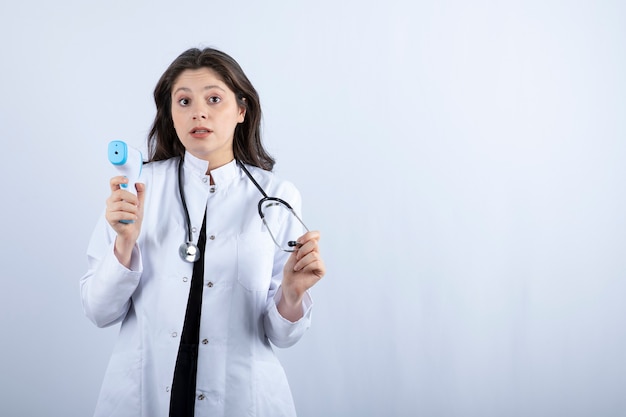 Portrait of female doctor showing thermometer and stethoscope on white wall.