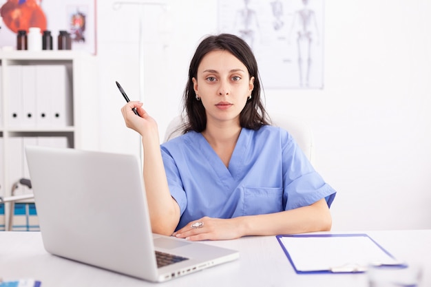 クリニックで彼女のラップトップコンピューターを使用している女性医師の肖像画。病院の職場でノートブックを使用している開業医、自信を持って、専門知識、医学。