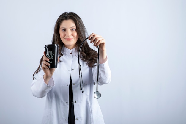 Portrait of female doctor with stethoscope and cup of coffee standing on white wall. 