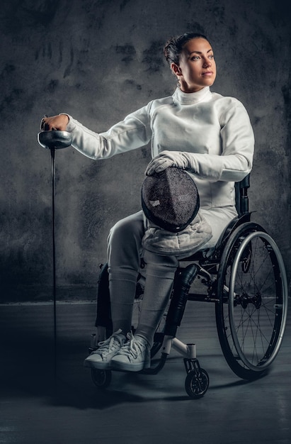 Portrait of female fencer in wheelchair with safety mask and rapier on grey background.