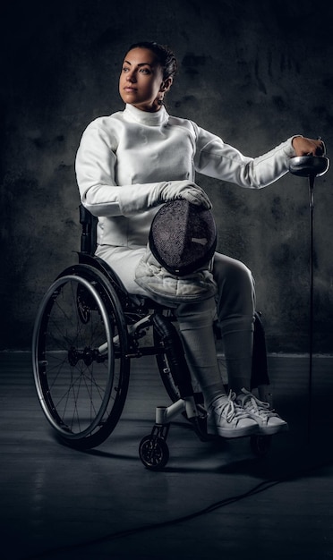 Portrait of female fencer in wheelchair with safety mask and rapier on grey background.