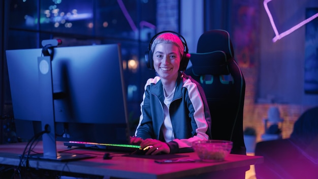 Portrait of a Female Gamer Playing Online Video Game on Computer Stylish Young Woman with Short Hair in Headphones Enjoying Leisure Time Smiling and Posing for Camera Cyber Gaming Neon Room