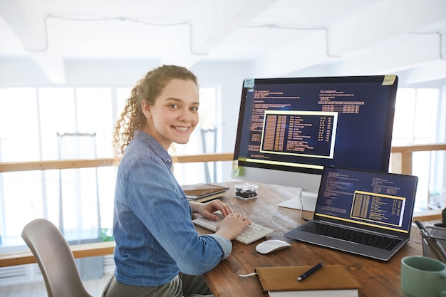 Portrait of female IT developer smiling at camera while typing on keyboard with black and orange programming code on computer screen and laptop in contemporary office interior, copy space