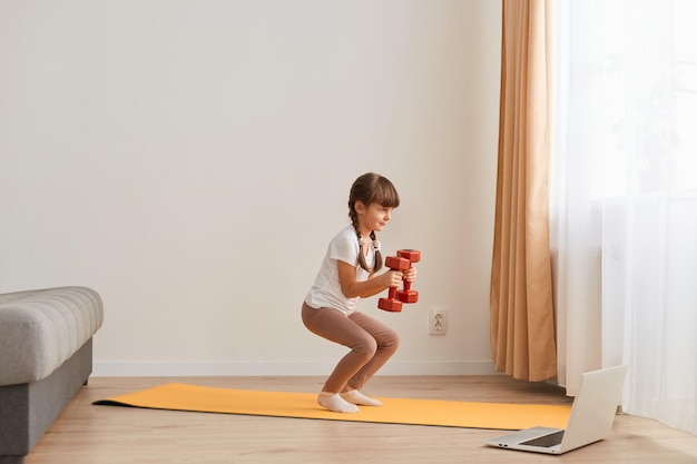 Portrait of female kid doing squatting workout at home cute kid training on a mat indoor darkhaired female kid having exercises with dumbbells in hands near the window