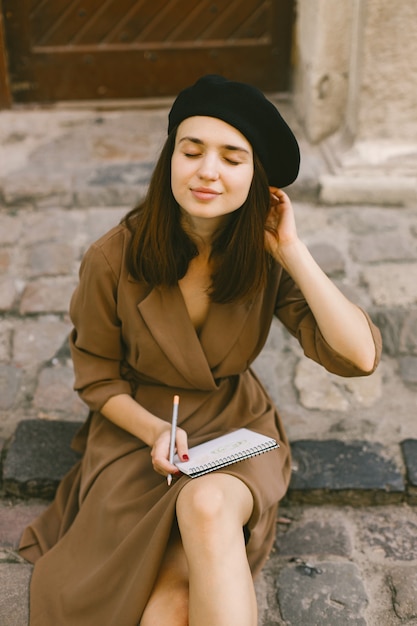 Portrait of a female painter painting while sitting in a city center. A woman wearing khaki dress and a black hat