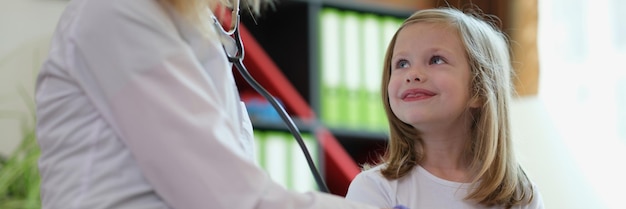 Portrait of female pediatrician examining little child with stethoscope smiling girl at doctor