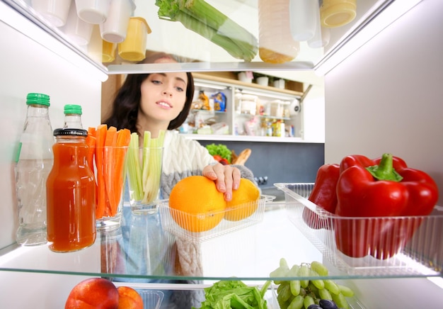 Портрет женщины, стоящей возле открытого холодильника, полного здоровых овощей и фруктов