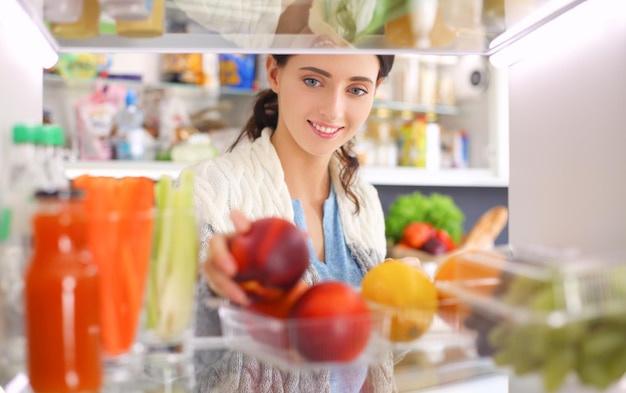 健康食品の野菜や果物でいっぱいの開いた冷蔵庫の近くに立っている女性の肖像画