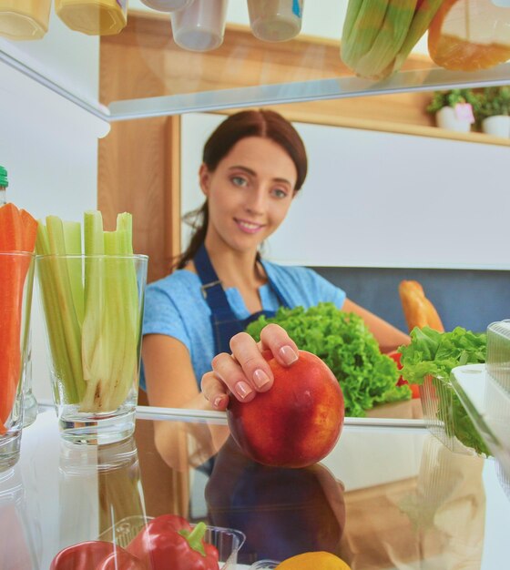 健康食品の野菜や果物の肖像画でいっぱいのオープン冷蔵庫の近くに立っている女性の肖像画