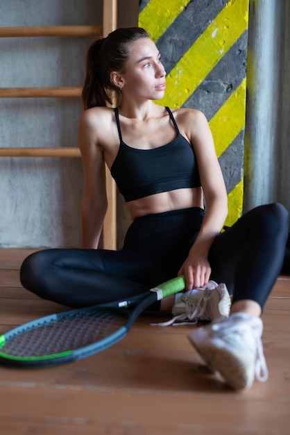 Portrait of female tennis player with racket sitting on gym floor