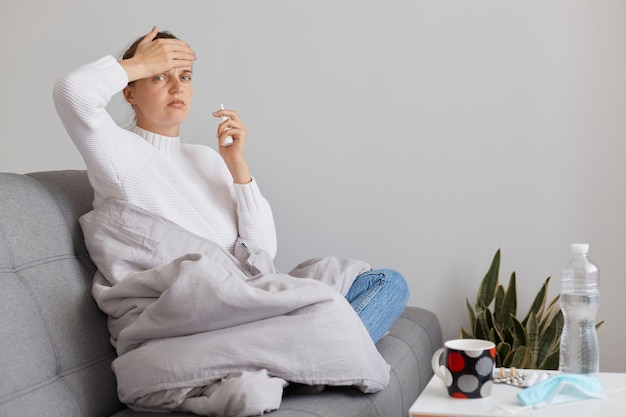 Portrait of female with headache and high temperature sitting on cough in plaid and looking at camera with sad expression grippe or catching cold symptoms