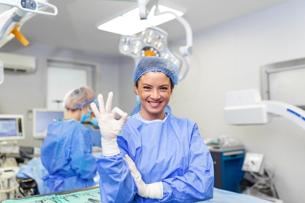 Portrait of female woman nurse surgeon OR staff member dressed in surgical scrubs gown mask and hair net in hospital operating room theater making eye contact smiling showing ok sign