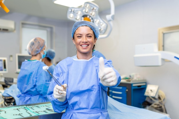 Portrait of female woman nurse surgeon or staff member dressed\
in surgical scrubs gown mask and hair net in hospital operating\
room theater making eye contact smiling thumbs up