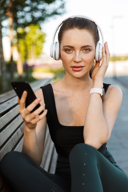 Portrait of feminine sportswoman wearing tracksuit holding smartphone and listening to music with headphones while sitting on bench in city park