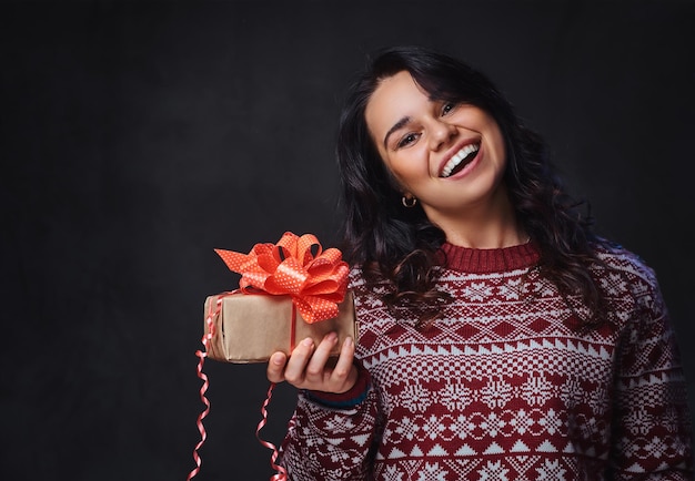Portrait of festive smiling brunette female with long curly hair, dressed in a red sweater holds Christmas gifts.
