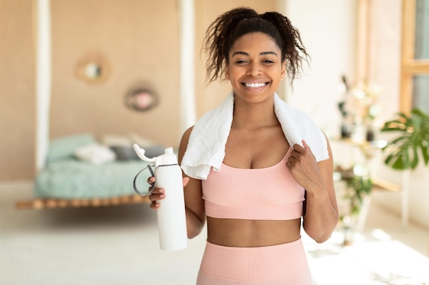 Portrait of fit black woman in sportswear and white towel on neck holding shaker with whey protein