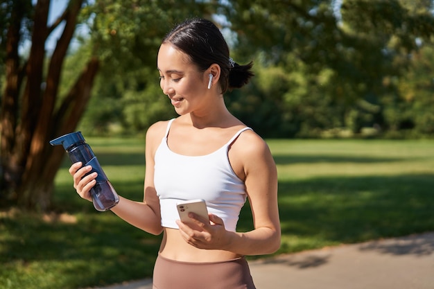 Portrait of fitness girl runner drinking water looking at smartphone standing in park in middle of w