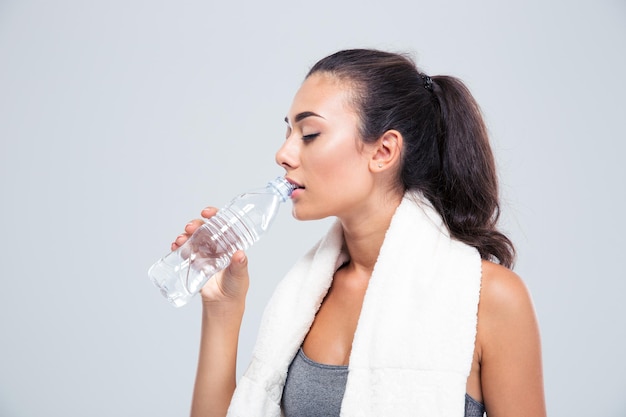 Portrait of a fitness woman with towel drinking water isolated on a white wall