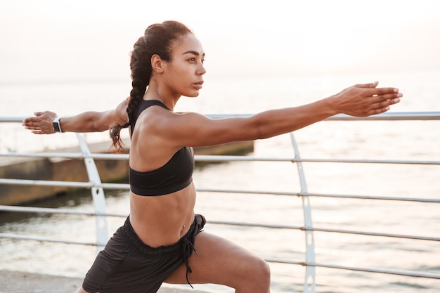 Portrait of fitness young woman in sportive clothes squatting and stretching her body while doing workout by seaside in morning