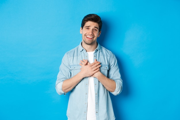 Portrait of flattered handsome man thanking you, holding hands on heart and smiling, looking grateful, standing over blue wall