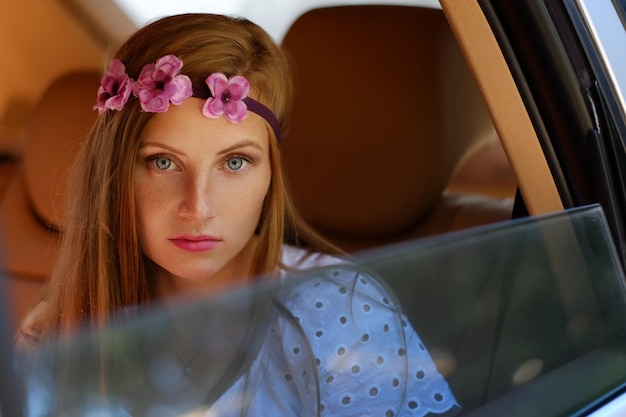 Portrait of flecked female with circle of flower on a head sits in a car on a back sit.