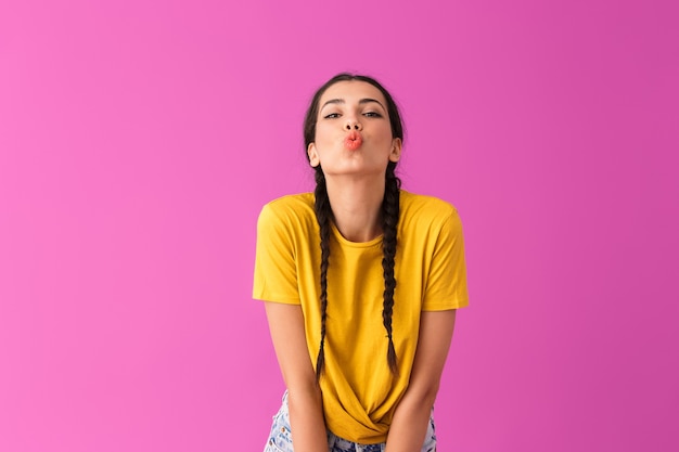 Портрет флиртующей молодой женщины в повседневной футболке, делающей поцелуй в губы, изолирован на розовой стене
