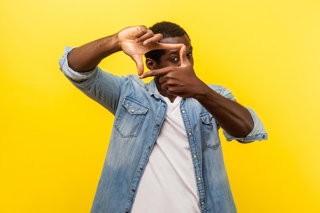 Portrait of focused curious man in denim casual shirt with rolled up sleeves looking through photo frame made of hands viewing distant with interest indoor studio shot isolated on yellow background