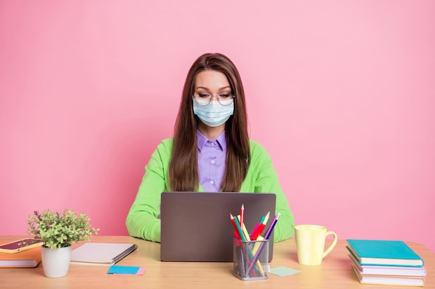 Ritratto di una ragazza concentrata che si siede al tavolo lavora a distanza, indossa un laptop indossa una camicia verde maschera medica isolato sfondo color pastello