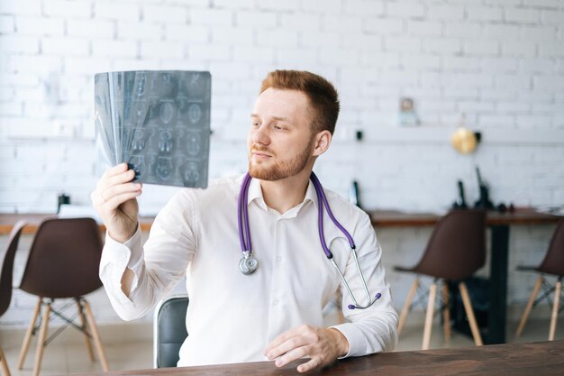 Portrait of focused male doctor wearing white uniform examining brain computerized tomography scan sitting at desk in hospital office, thinking about diagnosis. Concept of medicine and healthcare.