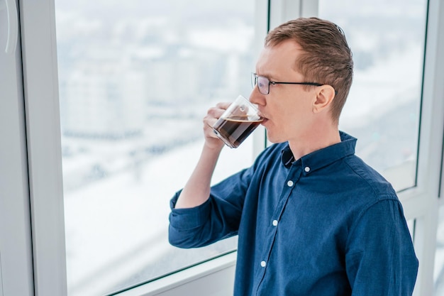 Portrait of focused middleaged thin man with short ginger hair glasses in blue longsleeve shirt looking out through window thoughtfully drinking black coffee from glass cup in morning on balcony