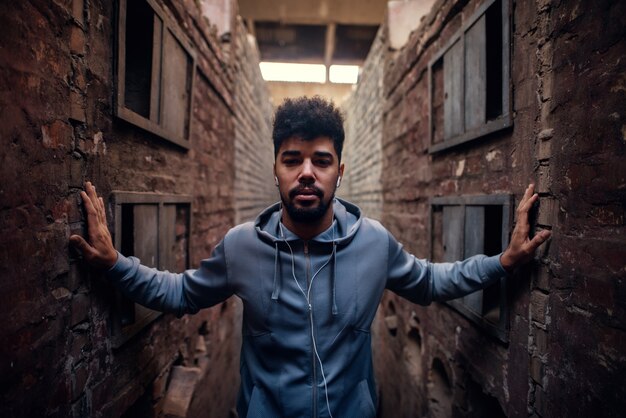 Photo portrait of focused motivated afro-american young attractive athletic man with earphones standing inside of the abandoned place in the middle of two walls