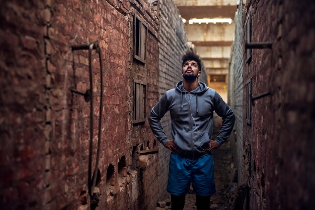 Portrait of focused motivated afro-american young handsome sportive man with earphones standing inside of the abandoned place in the middle of two walls and thinking while looking up.