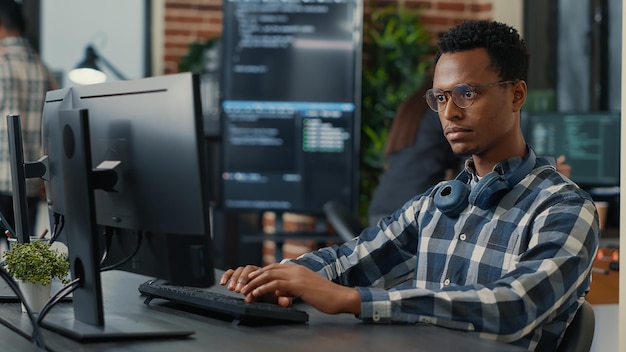 Portrait of focused programer writing code fixing glasses and\
smiling sitting at desk in it startup. coder programming user\
interface with team of programmers working in the background.