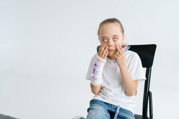 Portrait of fooling little girl drawing cute image with\
colorful marker on broken hand wrapped in white plaster bandage and\
showing whites of her eyes