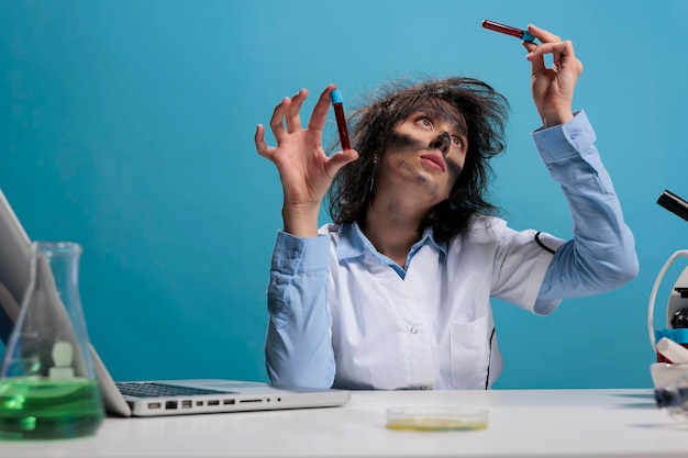 Portrait of foolish wild looking crazy chemist analyzing glass test tubes filled with blood sample while sitting at desk. Mad scientist with goofy aspect in laboratory examinating liquid substance.