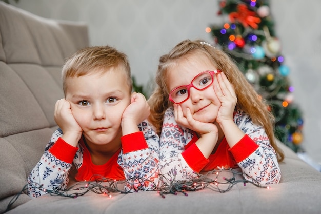 Ritratto di un fratello gemello di quattro anni e una sorella in pigiama a casa sul divano con l'albero di natale