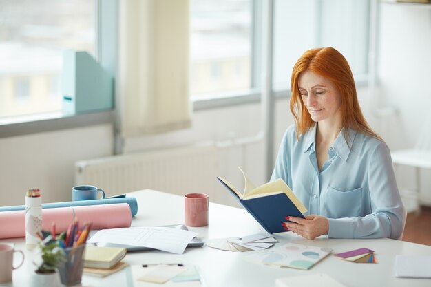 Portrait of freckled young woman reading journal while sitting at creative workplace by window, copy space