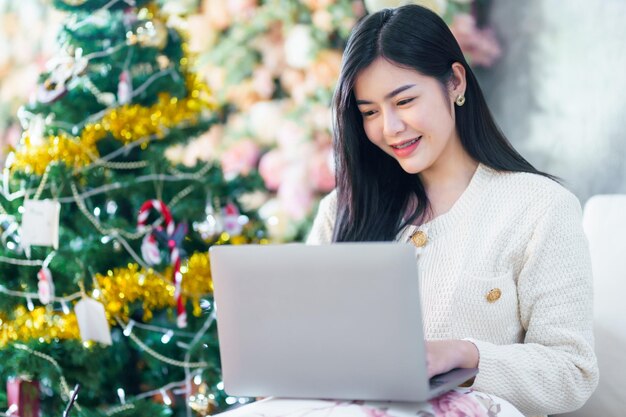 Ritratto freelance business bel sorriso positivo giovane donna asiatica online che lavora con il computer portatile a casa nel soggiorno al chiuso decorazione durante le vacanze di natale e capodanno.