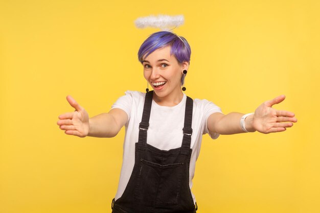 Photo portrait of friendly cute hipster girl with angelic halo above violet short hair and in denim overalls welcoming with raised hands and going to embrace isolated on yellow background studio shot