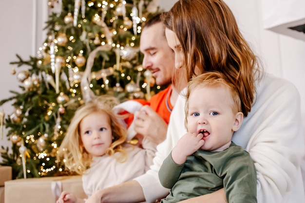 Portrait of friendly family are sitting at home near the Christmas tree, everyone is smiling. Focus on baby.