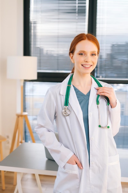 Portrait of friendly female doctor in white coat standing on background of window in sunny day in light medical clinic office.