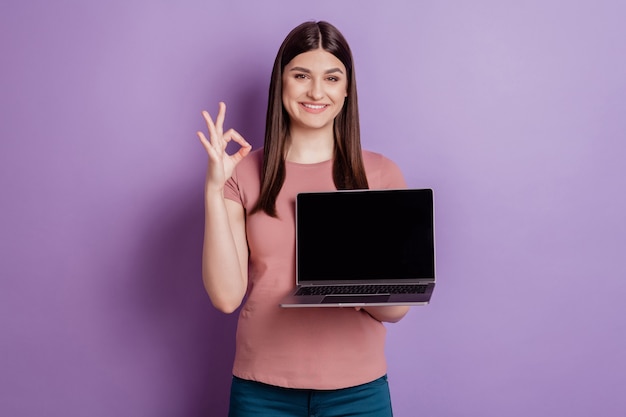 노트북 터치스크린을 들고 있는 친절한 소녀의 행복한 긍정적인 미소 손의 초상화는 보라색 배경에 격리된 괜찮은 광고를 보여줍니다.